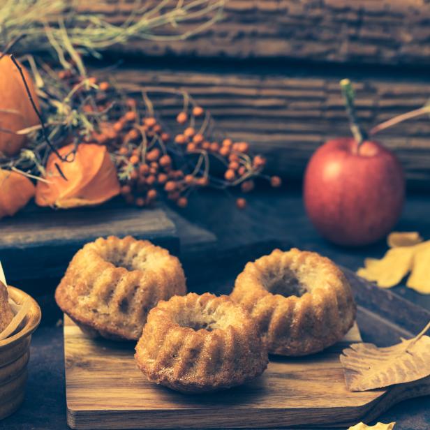 Ein Kürbis, Lampions, ein Apfel und kleine Gugelhupfe auf einem Holztisch.