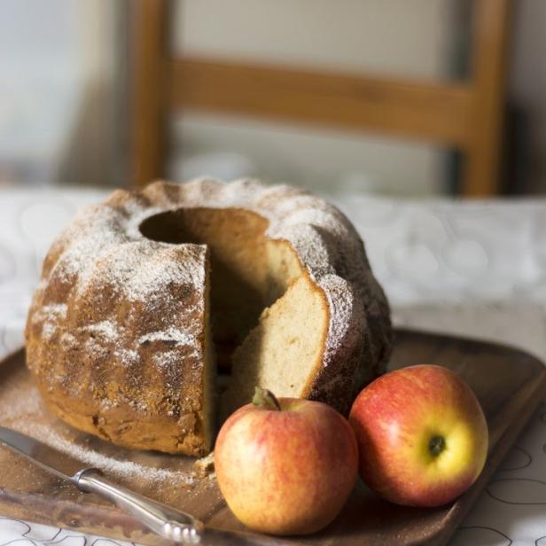 Ein angeschnittener Gugelhupf mit Puderzucker und zwei Äpfeln auf einem Holzbrett.