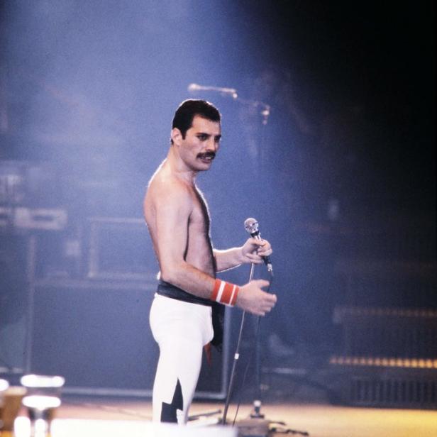 Freddie Mercury steht mit freiem Oberkörper und Mikrofon auf der Bühne.