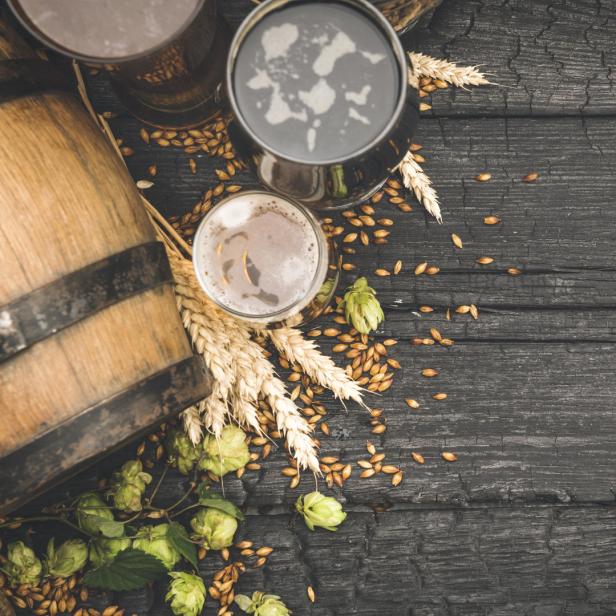 Drei Gläser Bier, Gerste, Hopfen und ein kleines Fass auf einem dunklen Holztisch.