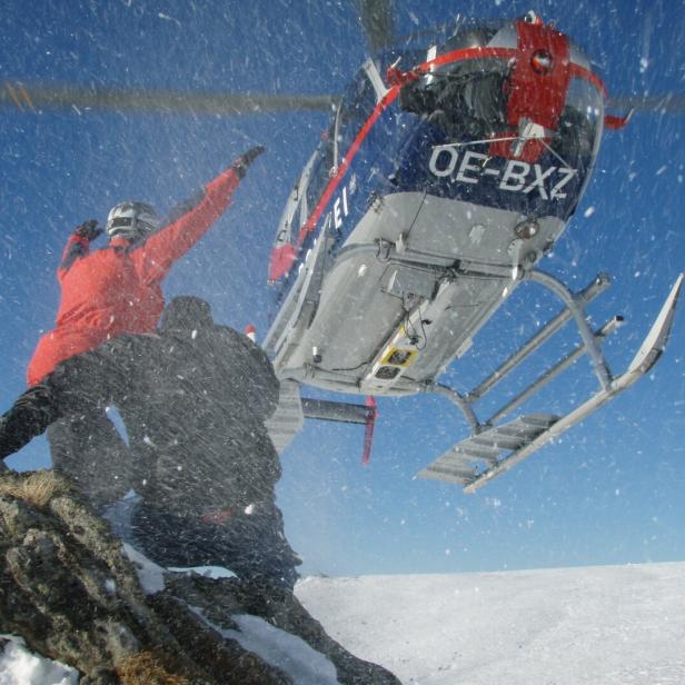 Ein Hubschrauber landet in einer verschneiten Berglandschaft, während zwei Personen in der Nähe stehen.