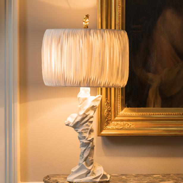 Eine beleuchtete Lampe mit weißem Schirm und skulpturalem Fuß steht auf einem Tisch.