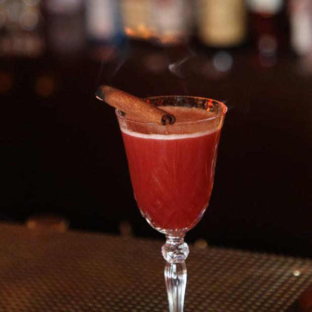 Ein roter Cocktail in einem Glas mit Zimtstange als Dekoration auf einer Bar.