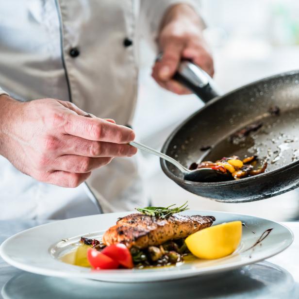 Ein Koch garniert ein Fischgericht mit Gemüse aus einer Bratpfanne.