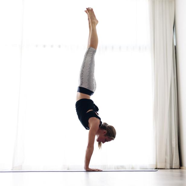 Eine Frau macht einen Handstand auf einer Yogamatte vor einem Fenster.