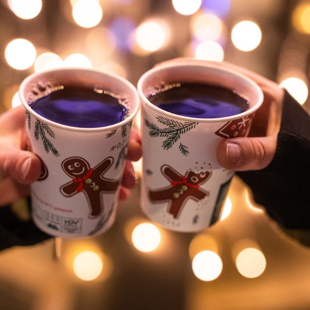 Zwei Hände halten Becher mit Glühwein vor einem Hintergrund mit Lichtern.