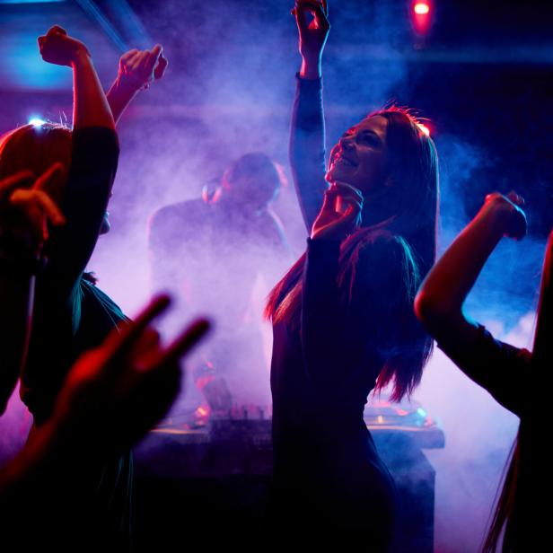 Menschen tanzen in einem Nachtclub mit farbiger Beleuchtung und einem DJ im Hintergrund.