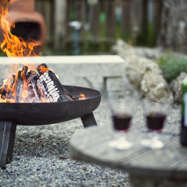 Ein Feuer brennt in einer Feuerschale, daneben stehen Wein und Gläser auf einem Tisch.