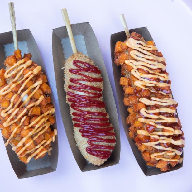 Drei koreanische Corn Dogs mit verschiedenen Toppings auf einem rosa Tablett.