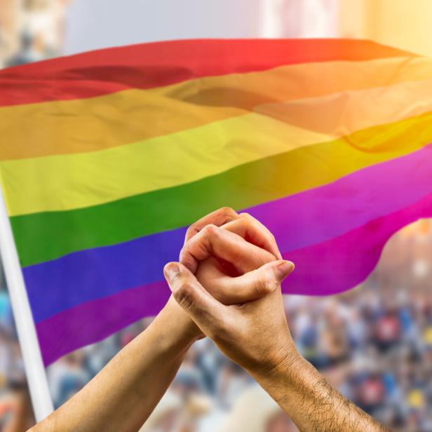 Zwei Hände halten sich vor einer Regenbogenflagge auf einer Demonstration.
