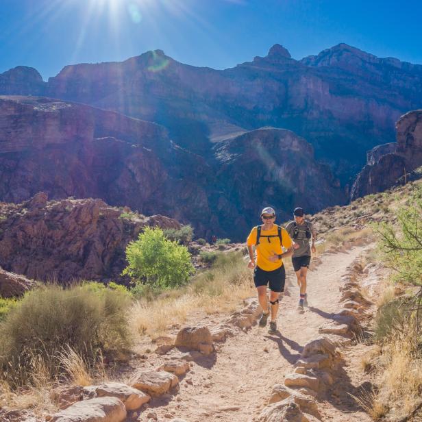 Zwei Männer joggen auf einem steinigen Pfad in einer bergigen Landschaft.