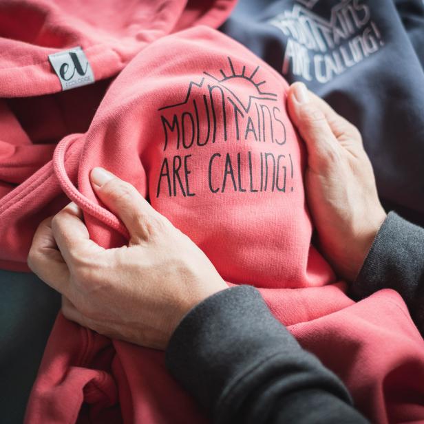 Hände halten einen rosa Hoodie mit der Aufschrift „Mountains are calling!“.