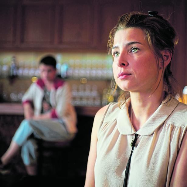 Eine Frau sitzt in einer Bar, im Hintergrund eine weitere Person.