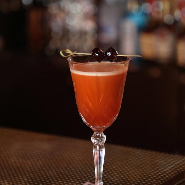 Ein Cocktail in einem eleganten Glas, garniert mit Kirschen, steht auf einer Bar.