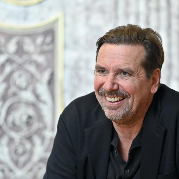Ein lächelnder Mann mit Schnurrbart und dunklem Sakko vor einer verzierten Wand.