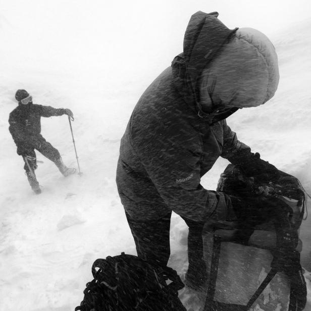 Bergsteiger packt seinen Rucksack im Schneesturm, während andere im Hintergrund zu sehen sind.