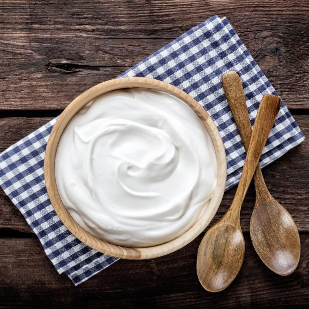 Eine Schale mit cremigem Joghurt steht auf einem karierten Tuch neben zwei Holzlöffeln auf Holztisch.