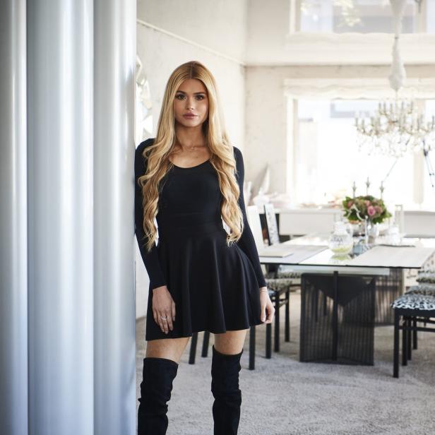 Eine Frau in einem schwarzen Kleid und Stiefeln posiert in einem hellen Esszimmer.