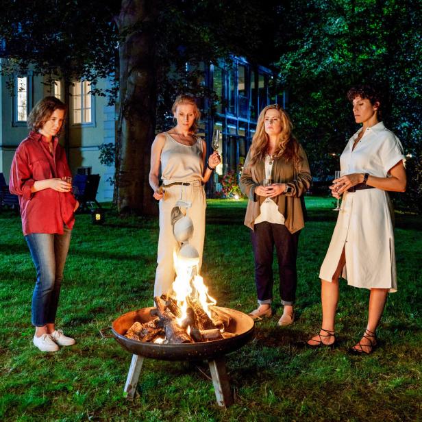 Vier Frauen stehen abends mit Getränken um eine Feuerschale im Garten.