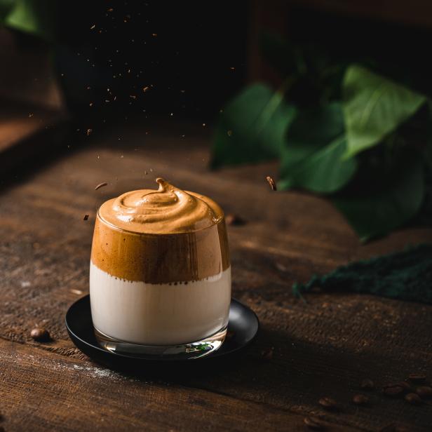 Ein Glas Dalgona-Kaffee steht auf einem Holztisch, Kaffeebohnen liegen daneben.