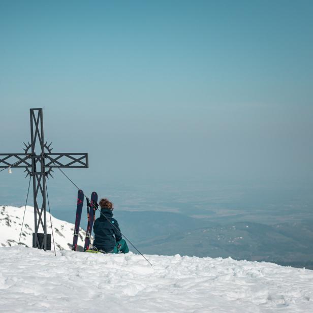 Eine Person sitzt neben einem Gipfelkreuz mit Skiern im Schnee.