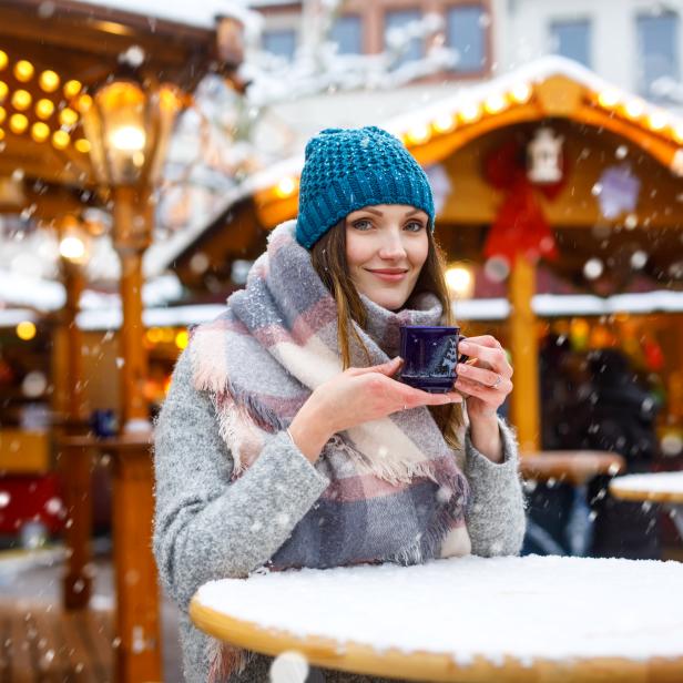 Eine Frau mit Mütze und Schal hält eine Tasse auf einem Weihnachtsmarkt im Schnee.