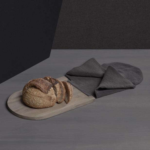 Ein angeschnittenes Brot liegt auf einem Holzbrett neben einer grauen Stoffserviette.