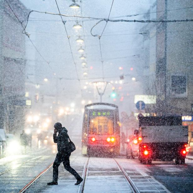 Eine Person überquert eine verschneite Straße mit einer Straßenbahn im Hintergrund.