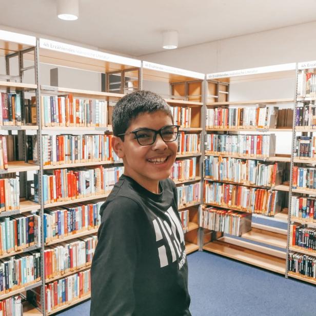 Ein lächelnder Junge steht vor Bücherregalen in einer Bibliothek.