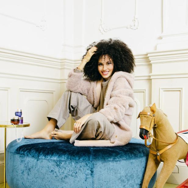 Eine Frau mit Afro sitzt barfuß auf einem blauen Samtpouf neben einem Schaukelpferd.