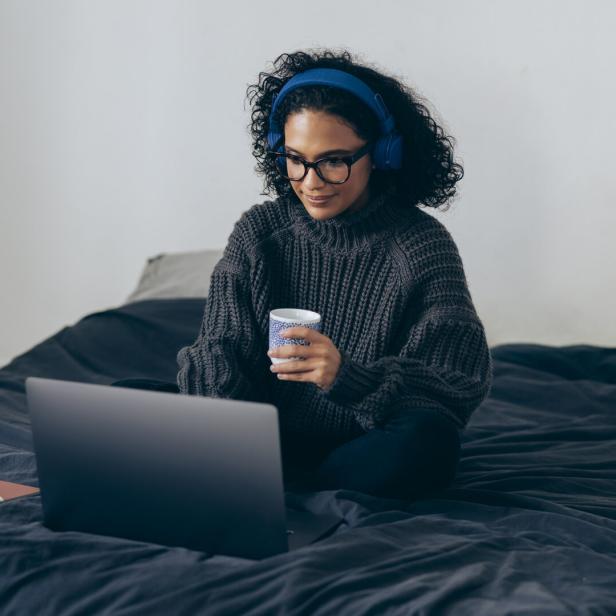 Eine Frau sitzt mit Kopfhörern auf dem Bett, arbeitet am Laptop und hält eine Tasse in der Hand.