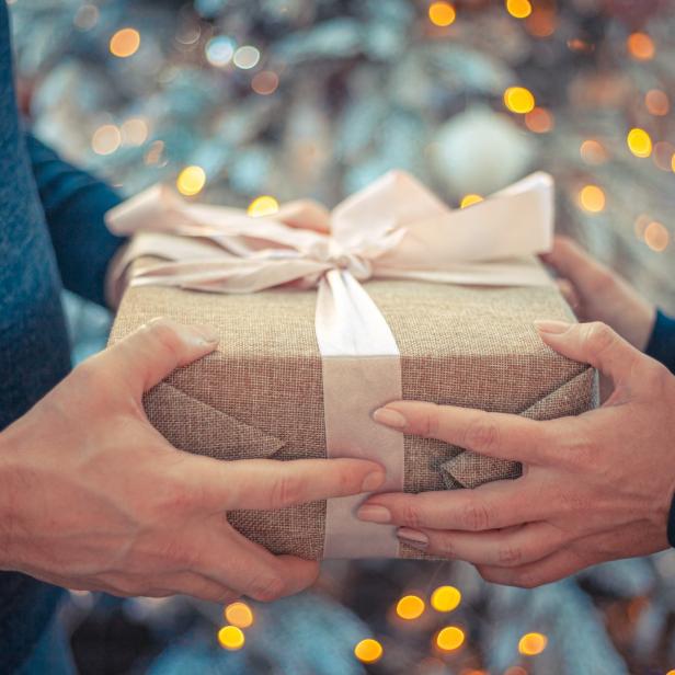 Zwei Hände halten ein Geschenk mit Schleife vor einem Weihnachtsbaum.