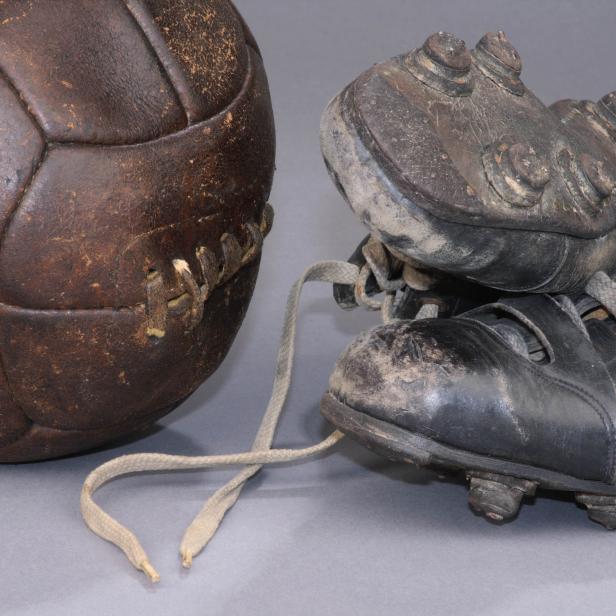 Ein alter, abgenutzter Fußball und ein Paar Fußballschuhe stehen auf einem grauen Untergrund.