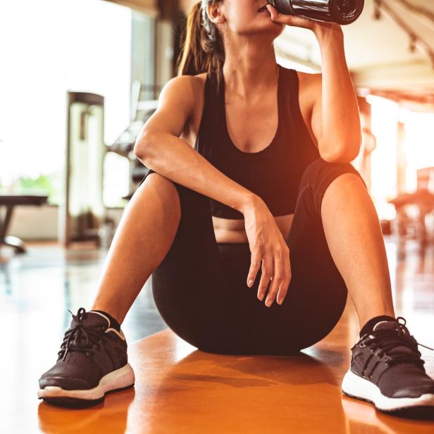 Eine Frau in Sportkleidung sitzt im Fitnessstudio und trinkt aus einer Flasche.