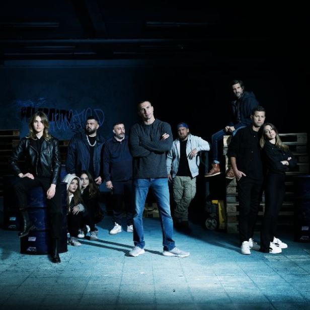 Eine Gruppe von zehn Personen posiert in einer Lagerhalle mit Graffiti an der Wand.