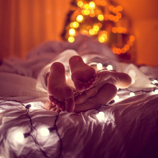 Füße ragen unter einer Decke hervor, im Hintergrund ein verschwommener Weihnachtsbaum und Lichterketten.