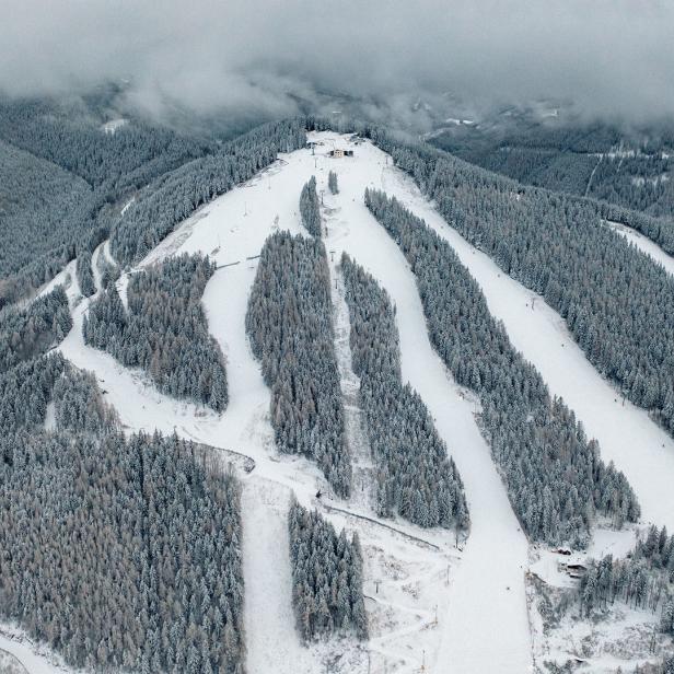 Luftaufnahme einer verschneiten Skipiste mit bewaldeten Hängen.
