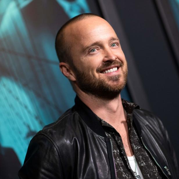 Porträt von Aaron Paul mit Bart und Lederjacke vor einem blauen Hintergrund.