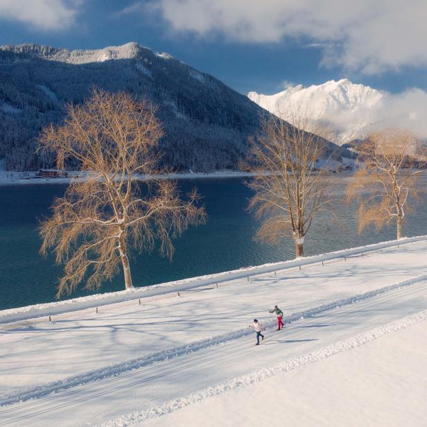 Zwei Personen laufen Ski auf einer schneebedeckten Loipe entlang eines Sees.