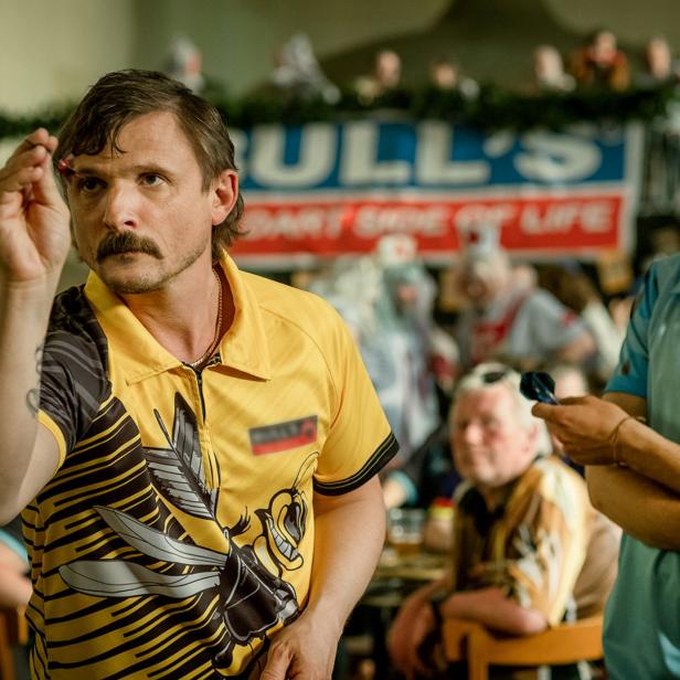 Ein Mann mit Schnurrbart zielt mit einem Dartpfeil auf eine Dartscheibe.