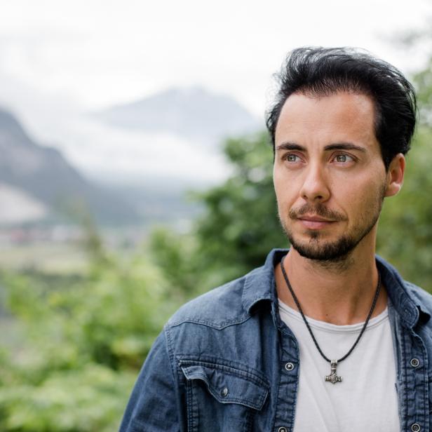Ein Mann mit Bart und Jeansjacke blickt vor einer Bergkulisse in die Ferne.