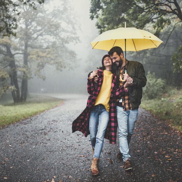 Ein Paar spaziert lachend mit einem gelben Regenschirm im Regen durch einen Park.