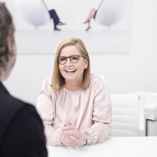 Eine lachende Frau mit Brille sitzt an einem Tisch und spricht mit einer Person.