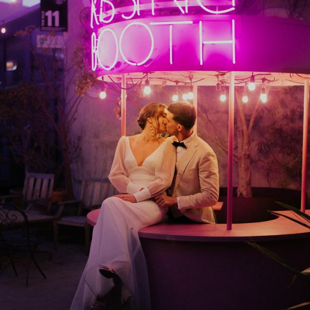 Ein Brautpaar küsst sich vor einem rosa „Kissing Booth“ mit Neonbeleuchtung.