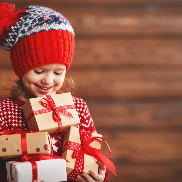 Ein lächelndes Mädchen mit Mütze hält mehrere Weihnachtsgeschenke im Arm.