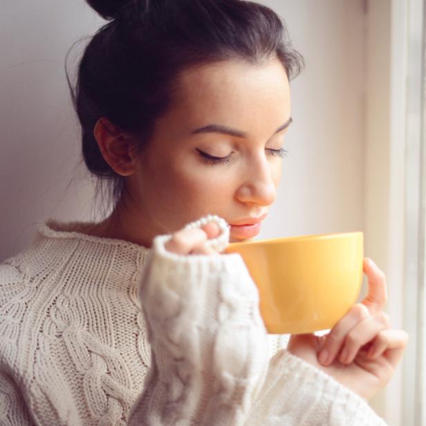 Eine Frau in einem weißen Pullover trinkt aus einer gelben Tasse am Fenster.