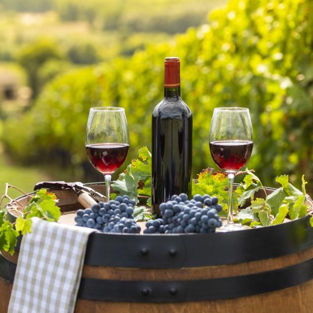 Eine Flasche Rotwein und zwei Gläser stehen auf einem Weinfass vor einem Weinberg.
