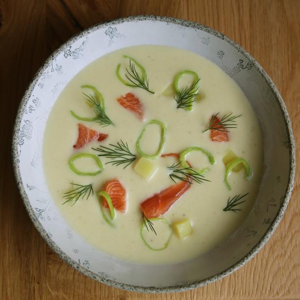 Eine Schüssel cremige Suppe mit Lachs, Kartoffeln und Dill auf einem Holztisch.