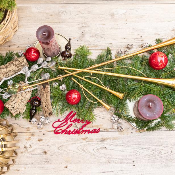 Weihnachtliche Dekoration mit Tannenzweigen, Kugeln, Kerzen und goldenen Trompeten auf einem Holztisch.