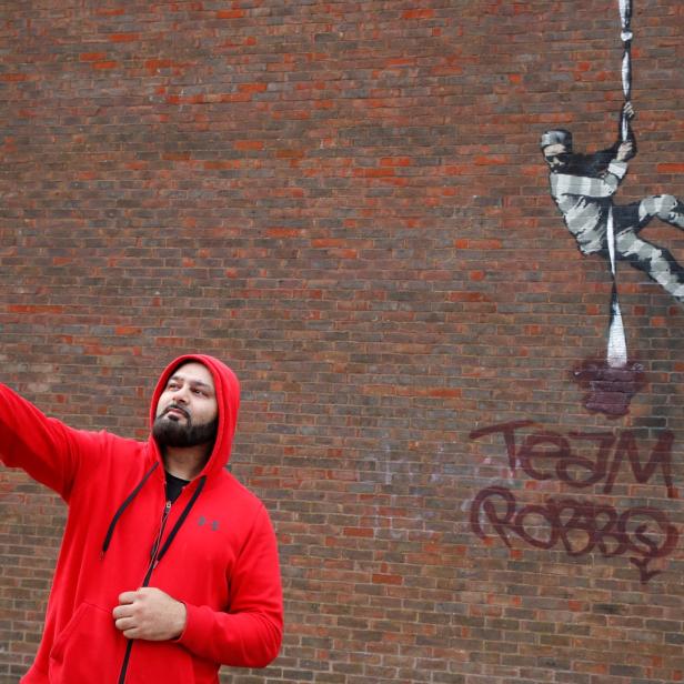 Ein Mann macht ein Selfie vor einer Backsteinmauer mit einem Banksy-Graffiti.